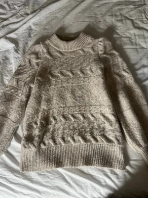 Beige kabelstickad tröja Vero Moda - Mysig beige kabelstickad tröja från Vero Moda med rund halsringning och ribbade muddar. Tröjan har ett klassiskt flätmönster och är perfekt för kyliga dagar. Materialet känns mjukt och skönt mot huden.