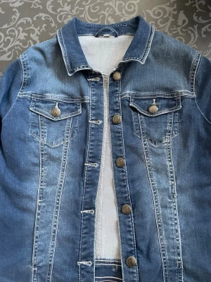 Blå jeansjacka  - Snygg blå jeansjacka från C•RO Jeans med klassisk krage, dekorativa bröstfickor med knappar och kontrastsömmar. Jackan har en rak passform och stängs med metallknappar framtill. Perfekt till alla dina outfits!