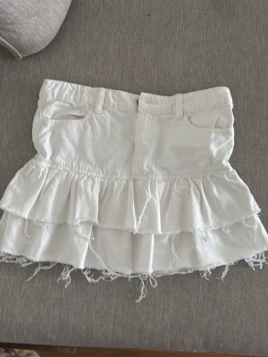 Vit volangkjol  - Snygg vit kortkjol med volanger och råa kanter. Kjolen har fickor fram och bak, bälteshällor och en cool, avslappnad look. Perfekt för dig som gillar en trendig och ungdomlig stil.