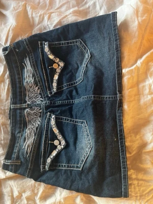 Mörk jeanskjol med broderade fickor - Cool mörkblå jeanskjol med två stora bakfickor, dekorerade med broderade vingar och detaljerade ficklock med knappar och mönstrad kant. Kjolen har markerade sömmar och en klassisk jeanslook som passar perfekt till sneakers eller boots. OBSERVERA defekt