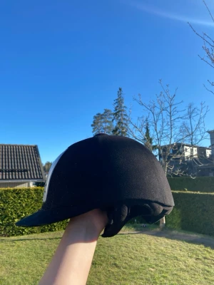 Svart ridhjälm med vit detalj - Säljer en svart ridhjälm med vit detalj på sidan. Hjälmen har en klassisk form och är vadderad på insidan för extra komfort. Storlek 56 (6 7/8). Perfekt för dig som rider och vill ha en stilren hjälm.