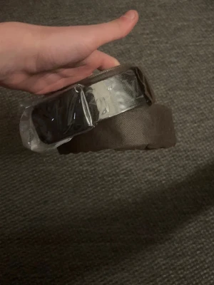Brunt tygbälte med silverfärgat spänne - Snyggt och stilrent bälte i brunt tyg med ett rektangulärt silverfärgat spänne. Perfekt för att ge en clean look till jeans eller chinos. Enkel design som passar till många outfits.