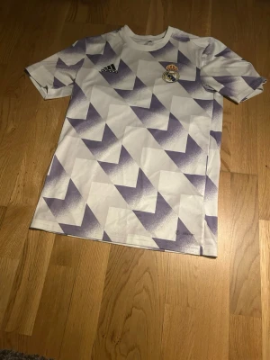 Real Madrid träningströja Adidas - Snygg Real Madrid träningströja från Adidas i vitt med lila geometriskt mönster. Tröjan har korta ärmar, rund hals och klubbens emblem på bröstet. Tillverkad i lätt och ventilerande material, perfekt för träning eller match.