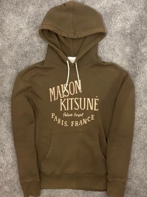Maison Kitsuné Hoodie - Snygg brun hoodie från Maison Kitsuné med vit text på bröstet och klassisk känguruficka. Huva med vita snören och mjukt material som känns skönt mot huden. Perfekt för en avslappnad och trendig look. Eftertraktad och är slut i varje lager online