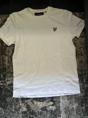 Vit t-shirt från Lyle & Scott - Vit t-shirt från Lyle & Scott med klassisk passform och korta ärmar. T-shirten har en liten gul broderad logga på bröstet och rund halsringning. Perfekt basplagg för garderoben.
