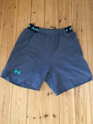Gråa träningsshorts från Under Armour - Snygga gråa träningsshorts från Under Armour med elastisk midja och turkost logotryck. Shortsen har meshfoder för extra ventilation och är perfekta för sport eller gym. Bekväm passform och sportig look.