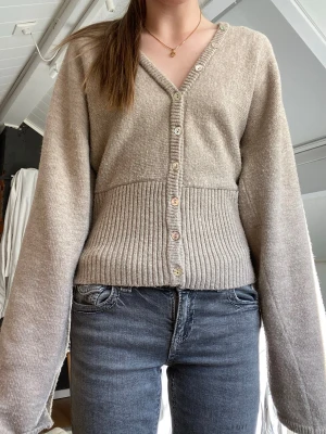 Beige kofta från Gina Tricot - Beige kofta med v-ringning från Gina Tricot. Koftan har ribbstickad midja och flerfärgade knappar framtill. Långärmad och i mjukt stickat material. 