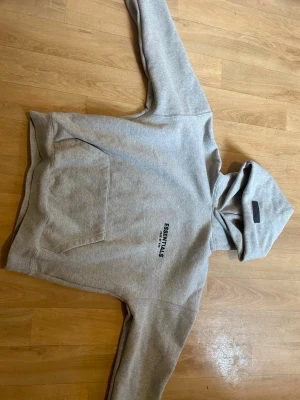 Grå Essentials hoodie med tryck - Snygg grå hoodie från Essentials med svart tryck på bröstet och patch på huvan. Klassisk känguruficka framtill och mjukt material som känns riktigt skönt. Perfekt för en avslappnad streetwear-look.