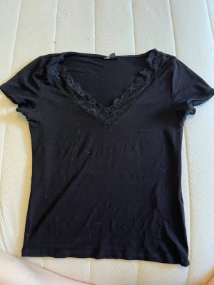 Svart topp med spetsdetalj - Svart topp med v-ringning och snygg spetsdetalj längs halsen. Kortärmad och figurnära passform, perfekt att matcha med jeans eller kjol. Materialet känns mjukt och stretchigt, troligen bomull eller polyester. Enkel och stilren design som passar till många outfits.
