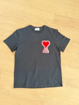 Svart t-shirt från Ami med hjärtdetalj - Svart t-shirt från Ami med en stor röd hjärtdetalj och bokstaven A på bröstet. Klassisk rund halsringning och korta ärmar. Perfekt för en trendig och avslappnad stil.