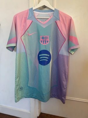 Pastellfärgad FC Barcelona tröja Nike - Säljer en unik FC Barcelona matchtröja i pastellfärger med Nike-logga och Spotify sponsortryck. Tröjan har korta ärmar, V-ringning och är tillverkad i ett lätt, ventilerande funktionsmaterial. Färgerna går i rosa, ljusblått, lila och mintgrönt med grafiska detaljer.
