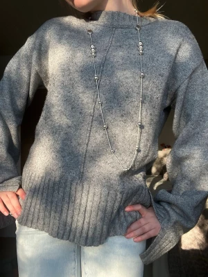 Grå oversized stickad tröja - Mysig grå stickad tröja med oversized passform och bred ribbstickad kant nedtill och vid ärmsluten. Tröjan har en hög hals och är tillverkad i ett mjukt material som passar perfekt för kyliga dagar.