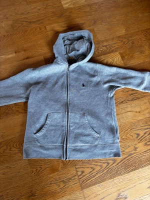 Grå hoodie från Polo Ralph Lauren - Grå zip-hoodie från Polo Ralph Lauren med klassisk svart broderad logga på bröstet. Hoodien är köpt på NK (stockholm) den har en liten skroma på armen (se bild 5) säljer nu pågrund av att den är liten, har använts några få gånger. 