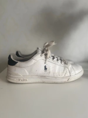 Vita Polo Ralph Lauren sneakers - Klassiska vita sneakers från Polo Ralph Lauren med mörkblå detalj på hälen och Polo-logga på sidan. Skorna har lågt skaft, vita snören och är tillverkade i läder. Perfekta för en clean och stilren look. Det är äkta och i bra skick kan behöva lätt tvätt men det inte några konstigheter och är enkelt köpte dom nya för 1300kr