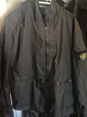 Svart vindjacka från Stone Island - Snygg svart vindjacka från Stone Island med klassisk patch på ärmen. Jackan har huva, dragkedja framtill och extra dragkedjor vid midjan. Tillverkad i lätt och slitstarkt material, perfekt för blåsiga dagar.