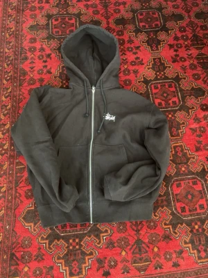 Svart Stüssy zip hoodie med logga - Svart hoodie med dragkedja från Stüssy. Stor vit Stüssy-logga på ryggen och mindre logga på bröstet. Klassisk känguruficka framtill och justerbar huva med snören. Perfekt streetwear-plagg i mjukt bomullsmaterial.