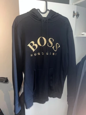 Hugo boss hoodie - Snygg svart hoodie från Hugo Boss med stor guldfärgad BOSS-logga på bröstet. Klassisk modell med huva och dragsnören samt en stor magficka framtill. Perfekt för en stilren och avslappnad look.