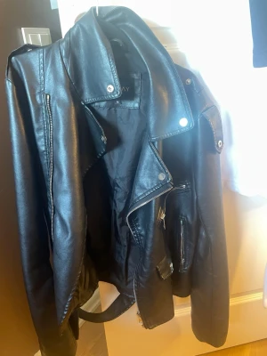 Svart skinnjacka från Missay - Snygg svart skinnjacka från Missay i klassisk bikerstil med dragkedjor, bred krage och detaljer i silver. Jackan har bälte i midjan och flera fickor med dragkedja. Perfekt för dig som vill ha en edgy look till din outfit.