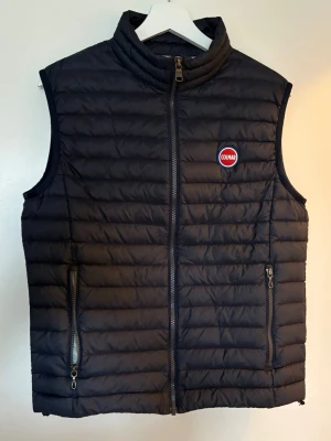Mörkblå dunväst från Colmar - Snygg svart dunväst från Colmar med quiltad design och hög krage. Västen har två dragkedjeförsedda fickor framtill och en diskret Colmar-logga på bröstet. Perfekt för lager-på-lager och håller dig varm utan att bli för tung.