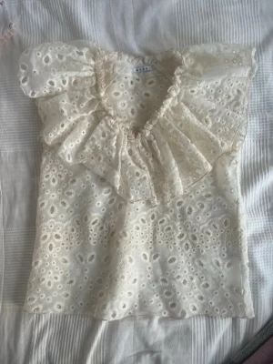 Vit broderad blus från Wera - Romantisk vit blus från Wera med broderade blommor och volangdetalj runt v-ringningen. Blusen är kortärmad och tillverkad i ett tunt, genombrutet tyg som ger en luftig känsla. Perfekt för en söt och trendig look.