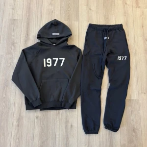Svart 1977-essentials tracksuit - Mysigt svart loungewear-set från Essentials med hoodie och matchande joggers. Båda plaggen har vit text '1977' tryckt på framsidan. Hoodien har en stor ficka och joggers har resår och snörning i midjan samt muddar vid benslut. Perfekt för chill dagar hemma. Passar dig som är 170cm+. Tar emot bud. Aldrig använd,tags kommer med.