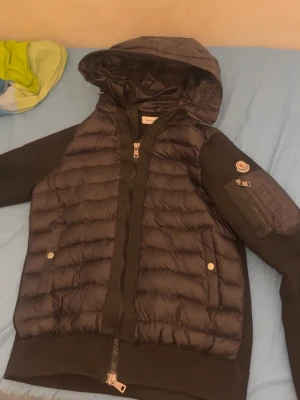 Svart dunjacka med huva från Moncler - Snygg svart dunjacka från Moncler med quiltad front och huva. Jackan har stickade ärmar, två fickor med knappar och en ärmficka med dragkedja och Moncler-logga. Perfekt för kalla vinterdagar och riktigt stilren look.