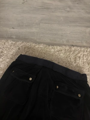 Svarta Juicy Couture mjukisbyxor - Säljer ett par mörkblåa  mjukisbyxor från Juicy Couture med bred resår i midjan och två bakfickor med guldfärgade knappar. Byxorna är i mjukt velourmaterial och har en klassisk, avslappnad passform.