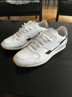 Vita sneakers från Oliver Cabell - Stilrena vita sneakers från Oliver Cabell med svarta detaljer på sidan och klassisk låg siluett. Skorna har vita snören, perforerade partier på tån och ovandelen är i slätt läder. Yttersulan är ljusbrun och ger en snygg kontrast. Storlek 44 och är i nyskick och endast testade,kommer med kartong och dustbag. Pris kan diskuteras. Hör av dig! 🤙 