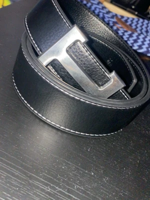 Svart skärp med H-spänne i skinn 105cm - Snyggt svart bälte i skinn med ett stort, silverfärgat H-format spänne. Bältet hakontrastsömmar längs kanterna som ger en clean och modern look. Perfekt accessoar för att lyfta vilken outfit som helst.