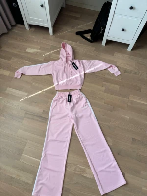 Rosa loungewear set från McKenzie - Snyggt rosa loungewear set från McKenzie med croppad hoodie och vida byxor. Setet har vita detaljer längs sidorna på byxorna och elastisk midja för skön passform. Perfekt för chill dagar hemma eller när du vill vara bekväm men ändå se fräsch ut.