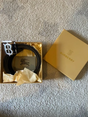 Burberry svart skinnbälte med silverdetalj 2 sidig - Burberry bälte i svart skinn med klassisk silverfärgad 'B'-spänne. Insidan har det ikoniska rutiga Burberry-mönstret i beige, rött och svart. Levereras i original ask med Burberry-logga och certifikat. 2 sidig