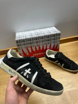 Premiata svarta sneakers med vit detalj - Svarta sneakers från Premiata med vit logotyp på sidan och vita detaljer vid hälen. Skorna har klassisk låg profil, svarta skosnören och en vit sula med mönster. Materialet är en kombination av mocka och läder för en stilren look.
