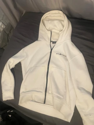 Vit zip-hoodie från Sail racing - Vit hoodie med dragkedja från Sail Racing. Klassisk modell med huva, två stora fickor framtill och svart logga på bröstet. Mjuk och skön, perfekt för chill dagar. Långärmad och i bomullsmix.