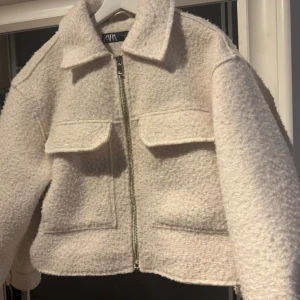 Vit teddyjacka med dragkedja - Säljer en vit teddyjacka med krage och två stora bröstfickor. Från Zara använd 1 gång, nypris 499kr. Jackan har en croppad passform och stängs med dragkedja framtill. Materialet är mjukt och fluffigt, perfekt för dig som gillar en chill och trendig look.