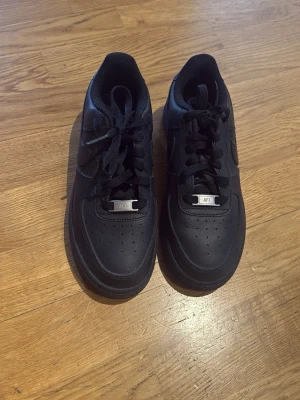 Nike Air Force 1 Low helsvarta sneakers - Säljer ett par klassiska Nike Air Force 1 Low sneakers i helsvart läder med perforerad tå och svarta skosnören. Snyggt och tidlöst design med metallplatta på snörningen där det står 'AF1'. Perfekta för dig som vill ha en clean och stilren look.