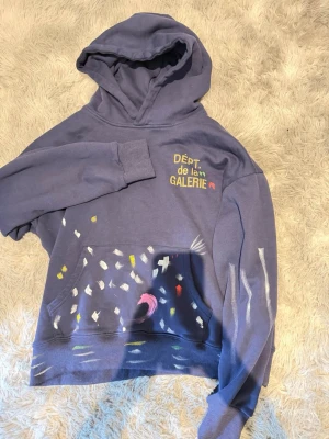 Blå hoodie från Dept de la Galerie/ Gallery dept - Cool blå hoodie med konstnärliga handmålade detaljer i vitt, gult, rosa och rött. Tryck på bröstet och unika penseldrag på både fram- och baksida samt ärmar. Mjuk bomullskänsla och klassisk huva. Perfekt för dig som gillar streetstyle och kreativ design.