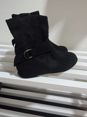 Svarta stövletter med spänne - Snygga svarta stövletter i mockaimitation med dekorativt spänne på sidan och dragkedja på insidan. Skorna har rundad tå och en något slouchy siluett som ger en avslappnad men trendig look. De är tunna och inte varma. De passar bra som inneskor vid finare tillfällen pga att de är så tunna. Endast använda vid ett kort tillfälle.