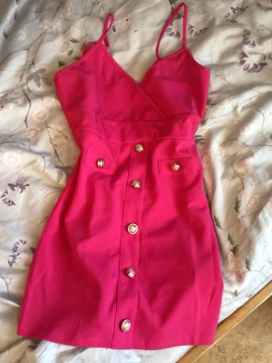 Kort rosa klänning med knappar - Snygg kort klänning i stark rosa färg med omlott upptill och smala axelband. Klänningen har dekorativa knappar framtill och två små fickor. Perfekt för dig som vill sticka ut med en färgglad look.