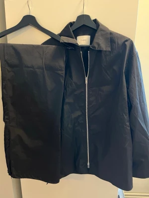 Set Svart jacka med dragkedja och byxor från CHIMI - Säljer en svart jacka från CHIMI med rak passform och silverfärgad dragkedja framtill. Jackan har klassisk krage och är tillverkad i ett lätt, glansigt material som ger en modern look. Byxorna har dragkedja nere för att kunna öppna upp. Perfekt för dig som gillar stilrena plagg.