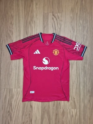 Manchester United 25/ 26 röd hemma matchtröja - Säljer en röd Manchester United matchtröja från Adidas med svarta detaljer och klubbens emblem på bröstet. Tröjan har korta ärmar, vit Snapdragon-logga på framsidan och svarta ränder på axlarna. Perfekt för fotbollsfans och sportaktiviteter.