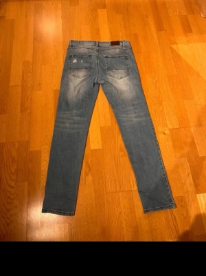 Blå slim fit jeans med slitningar - Säljer ett par blå slim fit jeans med diskreta slitningar och klassiska femficksdetaljer. Jeansen har en snygg tvätt med ljusare partier på låren och knäna. Perfekt för dig som gillar en modern och avslappnad look.
