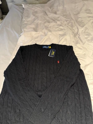Svart kabelstickad tröja Polo Ralph Lauren - Svart kabelstickad tröja från Polo Ralph Lauren med rund hals och lång ärm. Tröjan har ett klassiskt rött broderat logotyp på bröstet och ribbade muddar. Perfekt för dig som gillar stilrena och tidlösa plagg.