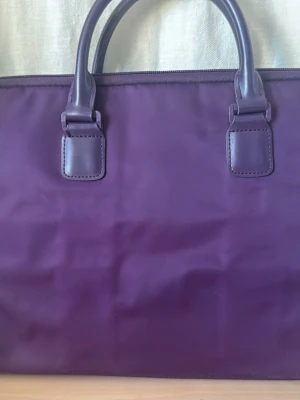 Lila datorväska med handtag och två fack - Snygg lila datorväska med två stadiga handtag och dragkedja. Väskan har ett stilrent och minimalistiskt utseende, perfekt för att bära din laptop. Materialet är slätt och syntetiskt, vilket gör den lätt att hålla ren. Två fack för enkel organisering. Mobilficka. Mkt sparsamt använd. 