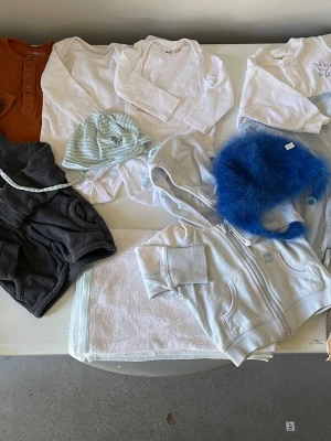 Startpaket babykläder och blöjor och våtservetter  - Säljer ett set med babykläder tio plagg och ett paket blöjor och våtservetter : två vita långärmade bodys, en brun långärmad body med knappar, en vit långärmad tröja med broderi, en grå luvjacka , en ljusblå randig mössa, en blå fluffig mössa, en bomulls filt och en ljusgrå zip-hoodie. Perfekt för nyfödd 