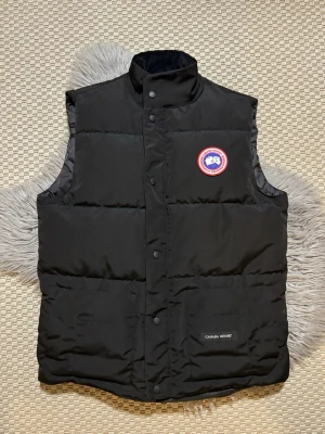Svart dunväst från Canada Goose - Snygg svart dunväst från Canada Goose med klassisk logga på bröstet och hög krage. Västen har tryckknappar framtill och två stora fickor, varav en med broderad logotyp. Perfekt för lager-på-lager och håller dig varm under kyliga dagar.