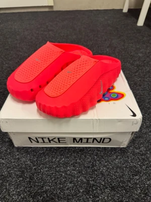 Nike Mind  - Säljer ett par Nike Mind tofflor i en stark röd färg med vågformad sula och ventilerande hål på sidorna. Tofflorna har en perforerad ovandel och silverfärgad Nike-logga på både ovansida och sida. Materialet är syntetiskt och designen är modern och unik. Stockx verifierade med tagg kvar