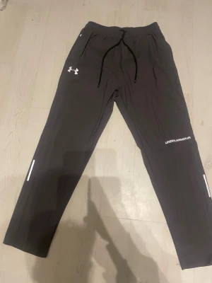 Svarta mjukisbyxor från Under Armour - Säljer ett par svarta mjukisbyxor från Under Armour med vita loggor på benet och höften. Byxorna har dragsko i midjan, raka ben och reflekterande detaljer vid anklarna. Perfekta för chill eller träning, riktigt sköna och stilrena.