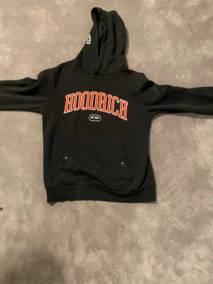 Svart hoodie från Hoodrich - Svart hoodie från Hoodrich med stor röd logotyp på bröstet och vita tryck på huvan. Hoodien har fickor med knappar framtill och klassisk huva. Materialet är mjukt och bekvämt, perfekt för en avslappnad streetwear-look.