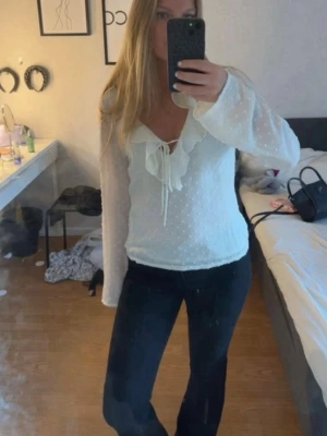 Vit blus med volang och knytband - Söt vit blus med diskret prickigt mönster, volangdetaljer längs ringningen och knytband framtill. Blusen har långa, lätt transparenta ärmar och en feminin, figurnära passform. Perfekt att styla med jeans eller kjol.