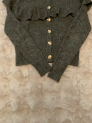 Grå kofta med volang från H&M - Mysig grå kofta från H&M med volangdetalj över bröstet och snygga guldfärgade knappar framtill. Långärmad och ribbstickad för extra komfort. Perfekt att slänga på sig när det är lite kyligt ute. Storlek XS , pris 30kr